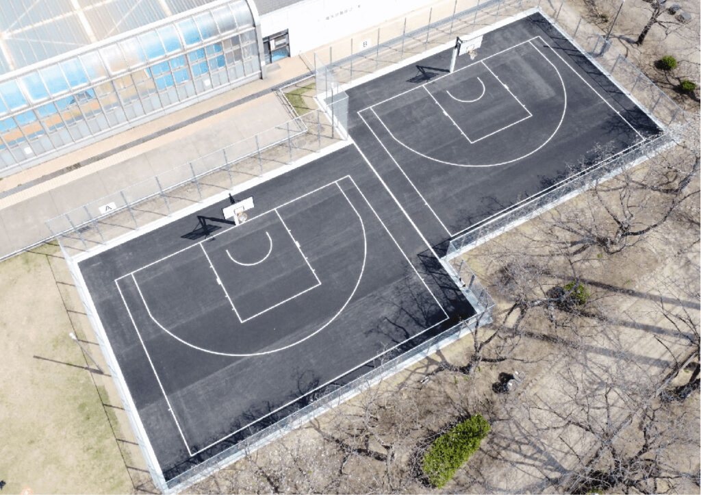 上空から撮影された南大分スポーツパークの3×3バスケットボールコートの写真。黒い舗装面に白線で区切られた2面のコートが並び、周囲は金属フェンスで囲まれている。左側には建物が隣接し、周囲には芝生や木々が配置された屋外スポーツエリアの様子が見える。