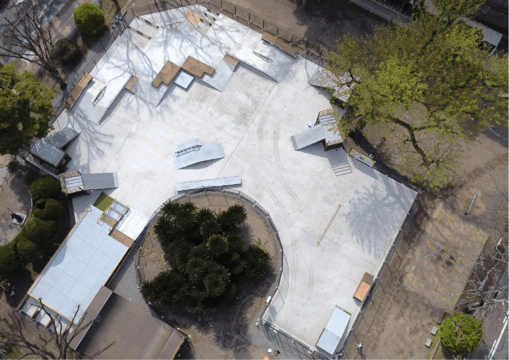 上空から撮影された大手公園スケートボードパークの写真。コンクリート製の広いスケートエリアに、ランプ、レール、段差などのセクションが配置され、中央には植栽のある円形スペースが設けられている。周囲はフェンスや木々に囲まれ、歩道やベンチが見える屋外スケートボード施設の全体レイアウトが俯瞰で捉えられている。