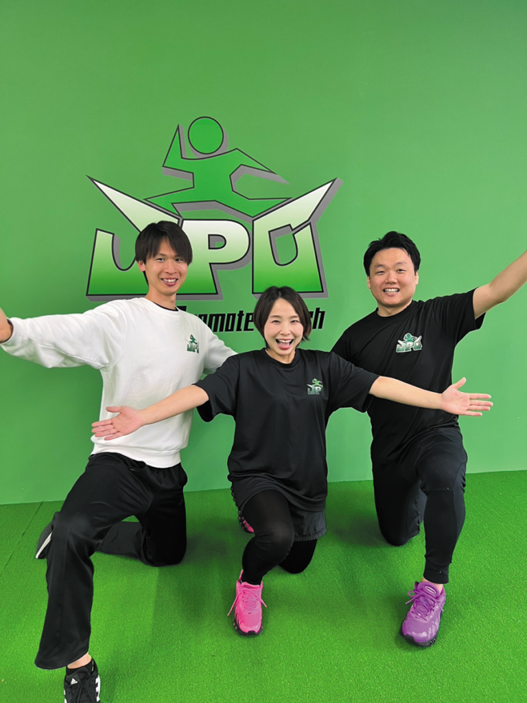 明るい緑色の壁を背景に、JPCスポーツ教室 大分古国府店のスタッフ3名がポーズをとっている写真。全員がスポーツウェアを着用し、胸元に同じロゴが入っている。片膝をつきながら腕を広げる動きでそろえ、背後にはロゴマークが掲げられた教室の雰囲気が伝わる構図。