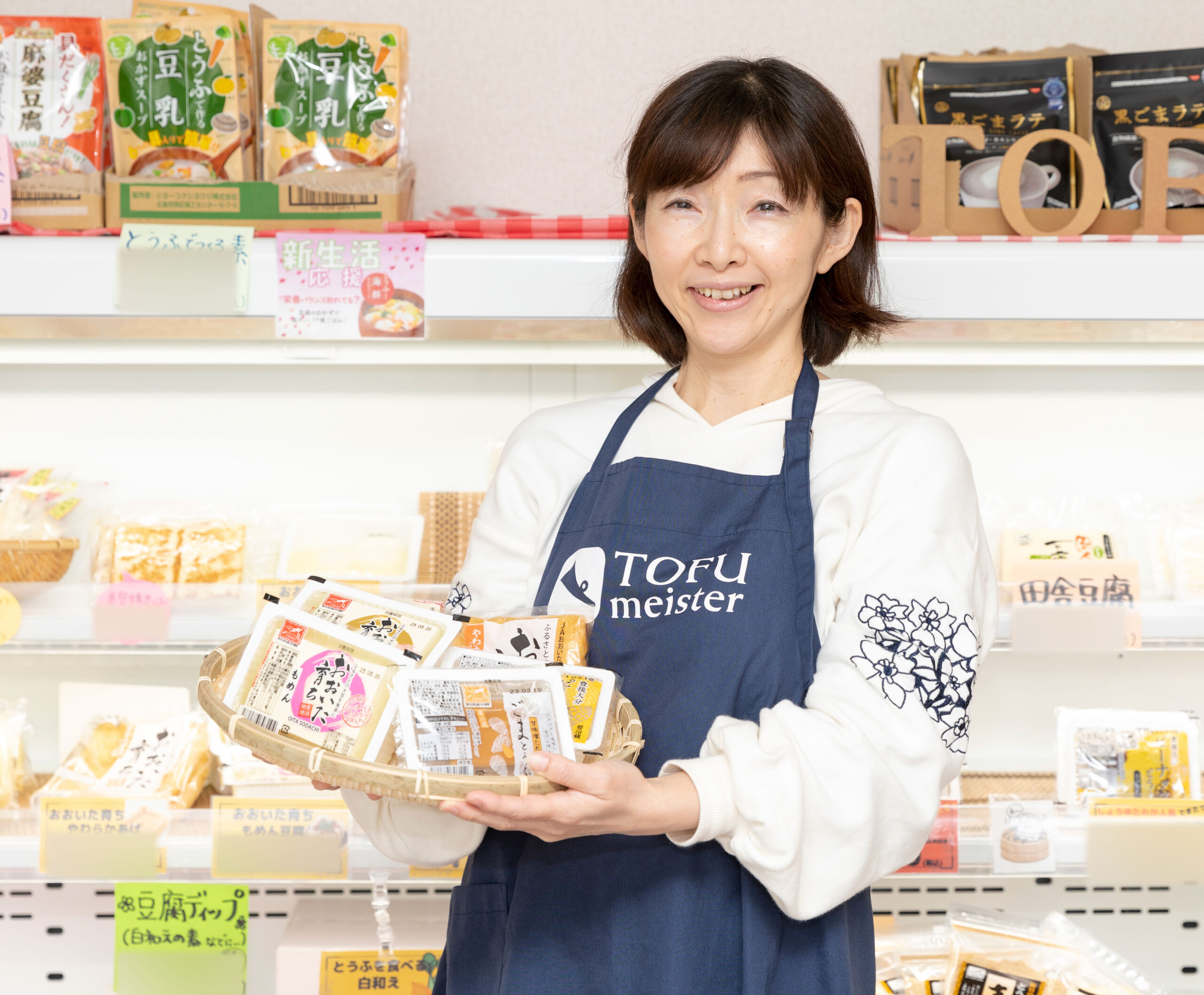 店内で豆腐製品を紹介する「合同会社 大豆のほいくえん大分」代表・大野こずえさんの写真。青いエプロンを身につけ、かごに豆腐や大豆加工品を入れて手に持っている。背後の棚には多様な豆腐商品が並び、明るい売り場の中で大野さんが笑顔で立つ姿が、大豆の魅力を伝える専門家としての雰囲気を表している。