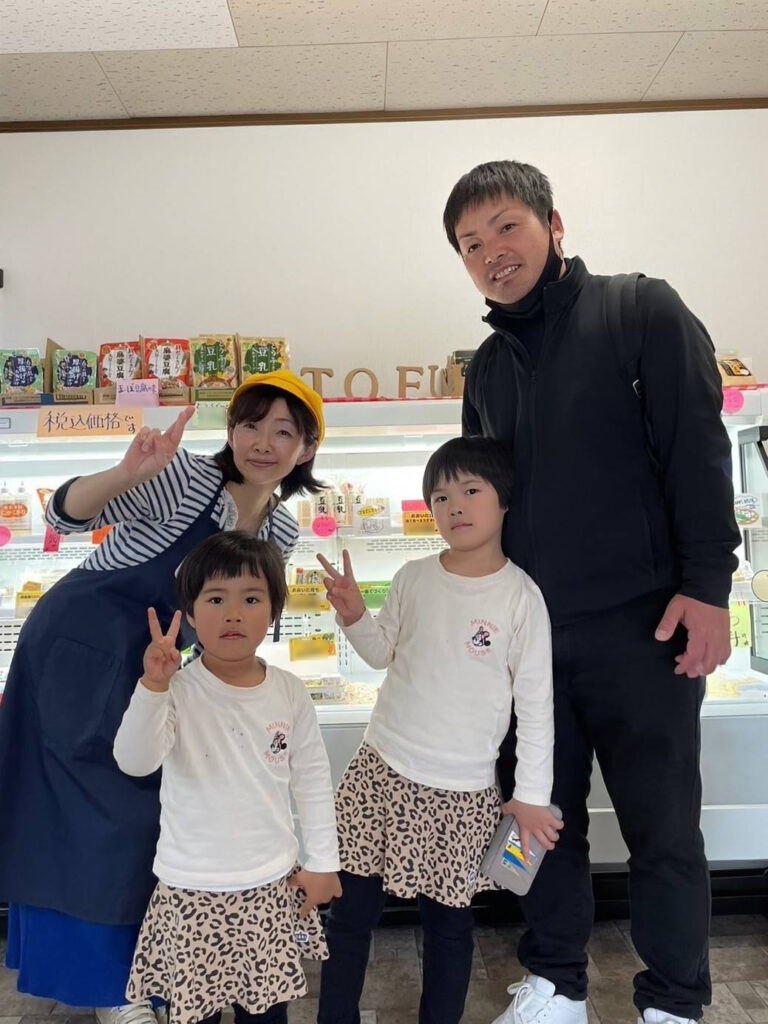 店内で豆腐製品が並ぶ冷蔵ケースの前に立つ4人の写真。青いエプロンと黄色い帽子を着用した人物が中央に立ち、その周囲に白いシャツと柄のスカートを着た2人の子ども、黒い服装の大人が並んでいる。背後の棚には豆腐や大豆加工品のパッケージが整然と並び、明るい売り場の雰囲気が伝わる。