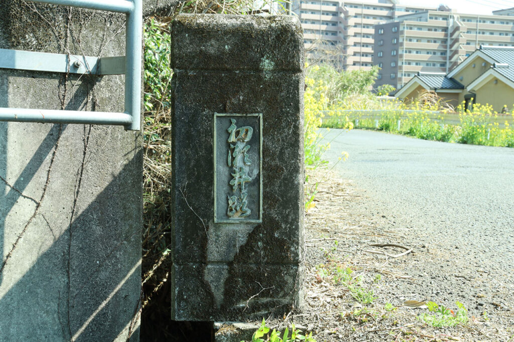 橋の欄干に取り付けられた「初瀬井路」と刻まれた金属製の橋名板を写した写真。コンクリート柱に黒地の銘板が固定され、横には金属製の手すりが続いている。周囲には黄色い花が咲く草地や道路、住宅が見える屋外の風景。