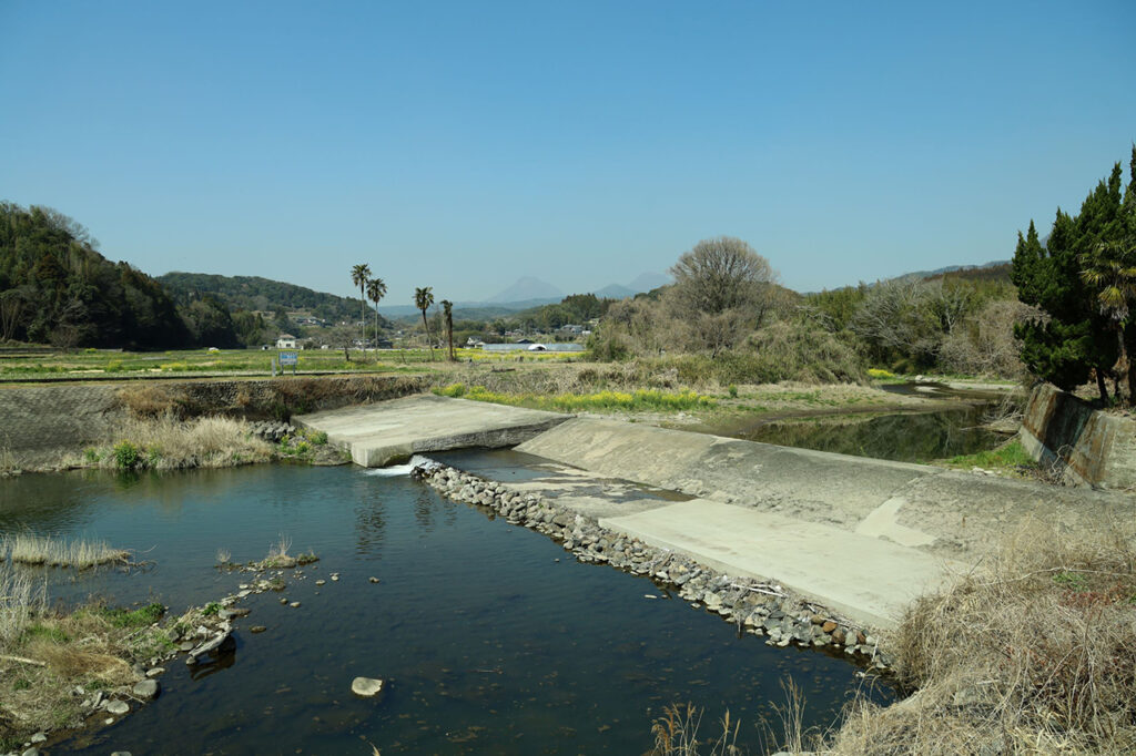 賀来川の水を堰き止める宮苑取入口を写した写真。浅い川にコンクリート製の堰が設けられ、水が段差を流れ落ちている。周囲には草地や低木が広がり、奥には農地や住宅、遠景に山並みが見える穏やかな田園風景。