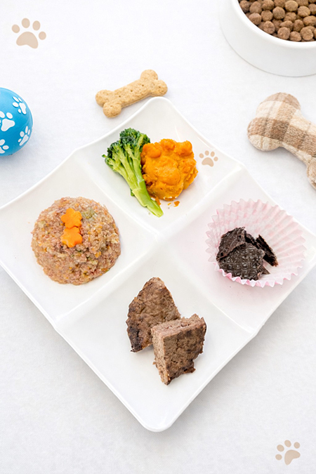 犬用ランチプレートが白い仕切り皿に盛り付けられた写真。ミックスフード、さつまいもとブロッコリー、肉の小片、カップに入ったトッピングなどが彩りよく配置されている。周囲にはドッグフードの入ったボウルやおもちゃ、骨型ビスケットが置かれ、ペット向けの食事シーンが演出されている。宗方倶樂部 トリミング＆カフェで提供される、わんこのランチプレートと飼い主向けランチの一部を紹介するイメージ。