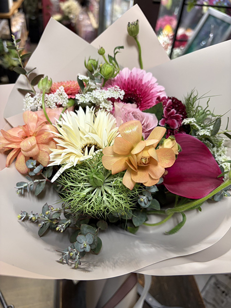 淡い色のラッピングに包まれた花束の写真。ピンク、オレンジ、クリーム、深い赤など複数の色合いの花が組み合わされ、グリーンがアクセントとして添えられている。STANDARD FLOWERのバースデー花束（3千円分）として、華やかで温かみのある色彩が特徴のアレンジメント。