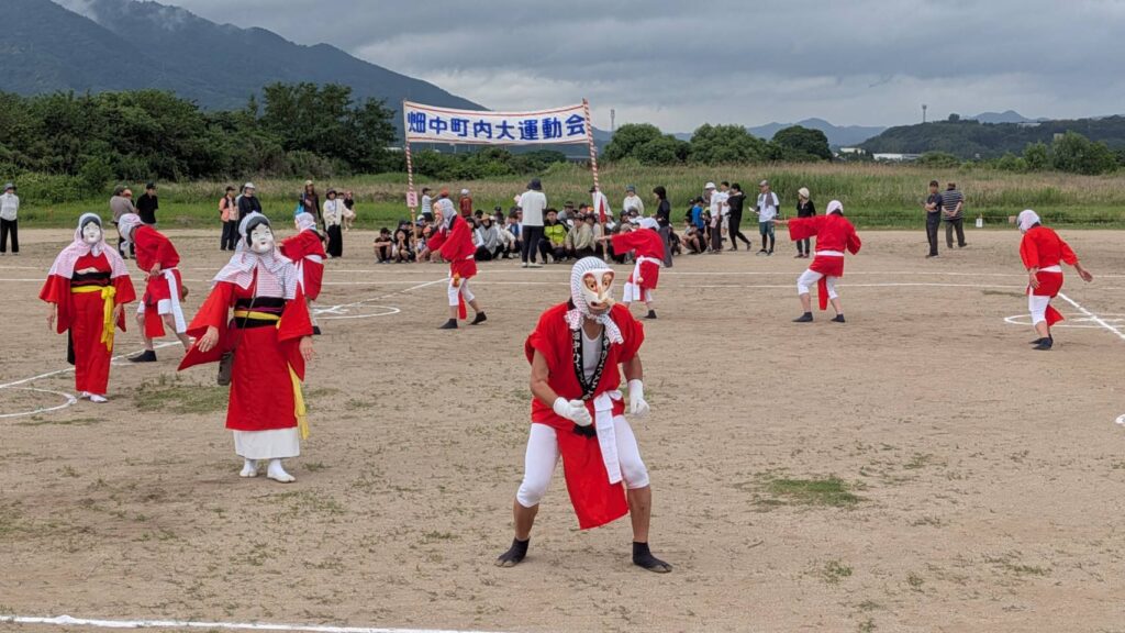運動場の中央で、赤い衣装と白い面をつけた出演者たちが並んでひょっとこ踊りをしている様子を写した写真。地面には白線が引かれ、背景には観客席や「畑中町内大運動会」の横断幕、山並みや木々が見える。地域の運動会で披露された伝統的な演目の雰囲気が伝わる構図。