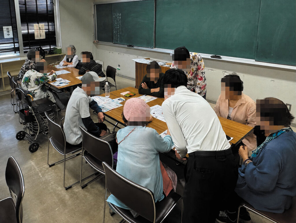 教室内で参加者たちがテーブルを囲み、資料を見ながら話し合いやワークに取り組んでいる様子を写した写真。講師が立って説明したり、参加者に寄り添ってサポートしたりする姿が見える。黒板や窓、歩行器などが置かれた室内で、「人生の最終章を自分で描く地図講座」の実際の学びの場が伝わる構図。