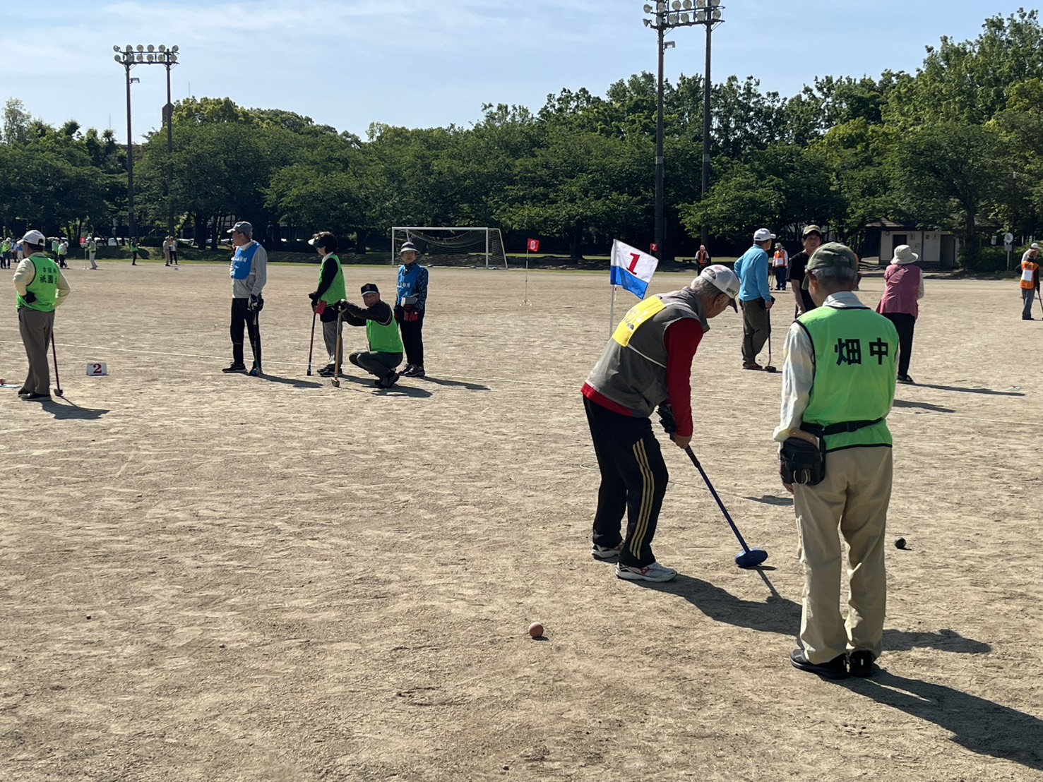 南大分スポーツパークのグラウンドで、参加者たちがクラブを構えてプレーしている様子を写した写真。色分けされたビブスを着た選手がボールを狙い、近くには番号旗やマーカーが置かれている。奥には他の参加者やゴールポスト、木々が見え、南大分体育協会主催の春季グラウンドゴルフ大会のにぎわいが伝わる構図。
