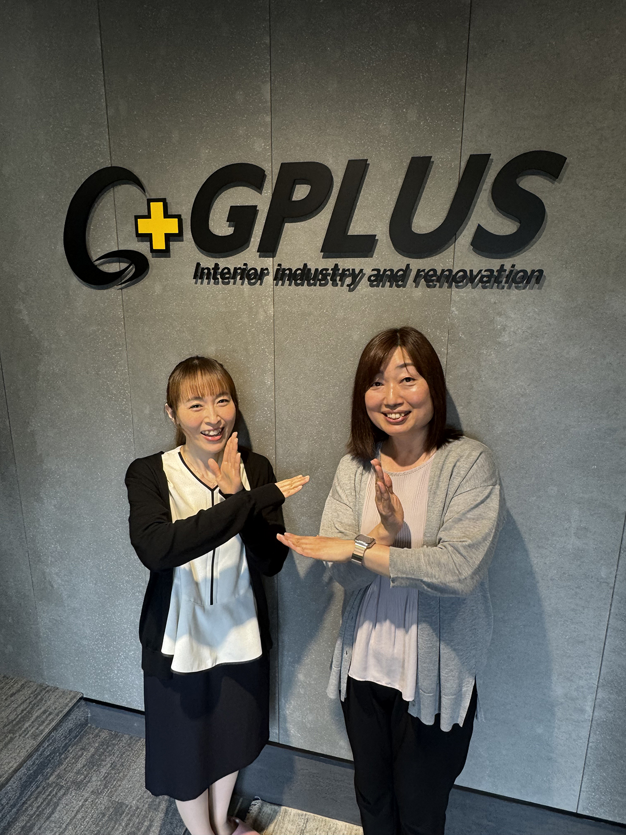GPLUSのロゴが掲げられた壁の前で、麻生さん（左）と安藤さん（右）が並んで立ち、腕で「プラス」のポーズをしている写真。グレー調の室内空間で、足元にはカーペットが敷かれ、企業の雰囲気が伝わるシンプルな構図。