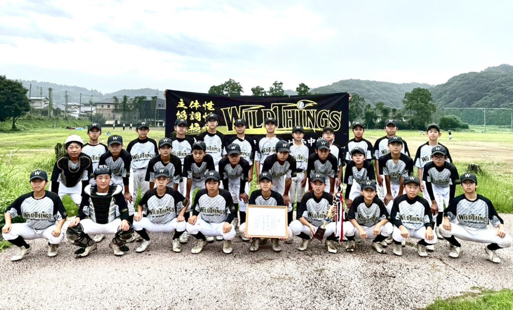 白いパンツと黒×グレーのユニフォームを着たBC南大分ワイルドシングスの選手たちが、屋外で横断幕を背に整列して記念撮影している写真。前列の選手が賞状やトロフィーを掲げており、チームの活躍を祝う雰囲気が伝わる構図。背景には芝生や丘が広がり、少年野球チームらしい爽やかな場面となっている。
