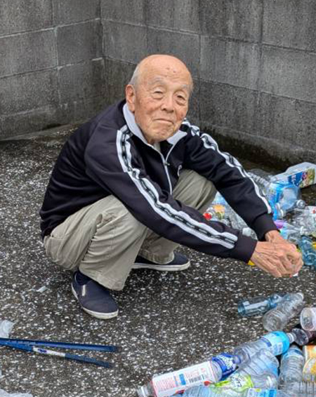 地面にしゃがみ込み、周囲に散らばったペットボトルや小さなごみを丁寧に集めている畑中在住の酒井潔さん（83歳）の様子を写した写真。コンクリートの壁際で作業する姿から、地域の環境美化に取り組む落ち着いた雰囲気が伝わる構図。