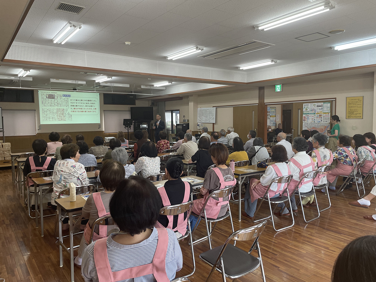 室内のスクリーン前で講師が説明を行い、テーブル席の参加者たちが資料を見ながら話を聞いている様子を写した写真。多くの参加者がピンク色のエプロンを着用し、木の床と明るい照明の会場に、地域サロン畑中での「墓じまい」勉強会の落ち着いた雰囲気が伝わる構図。