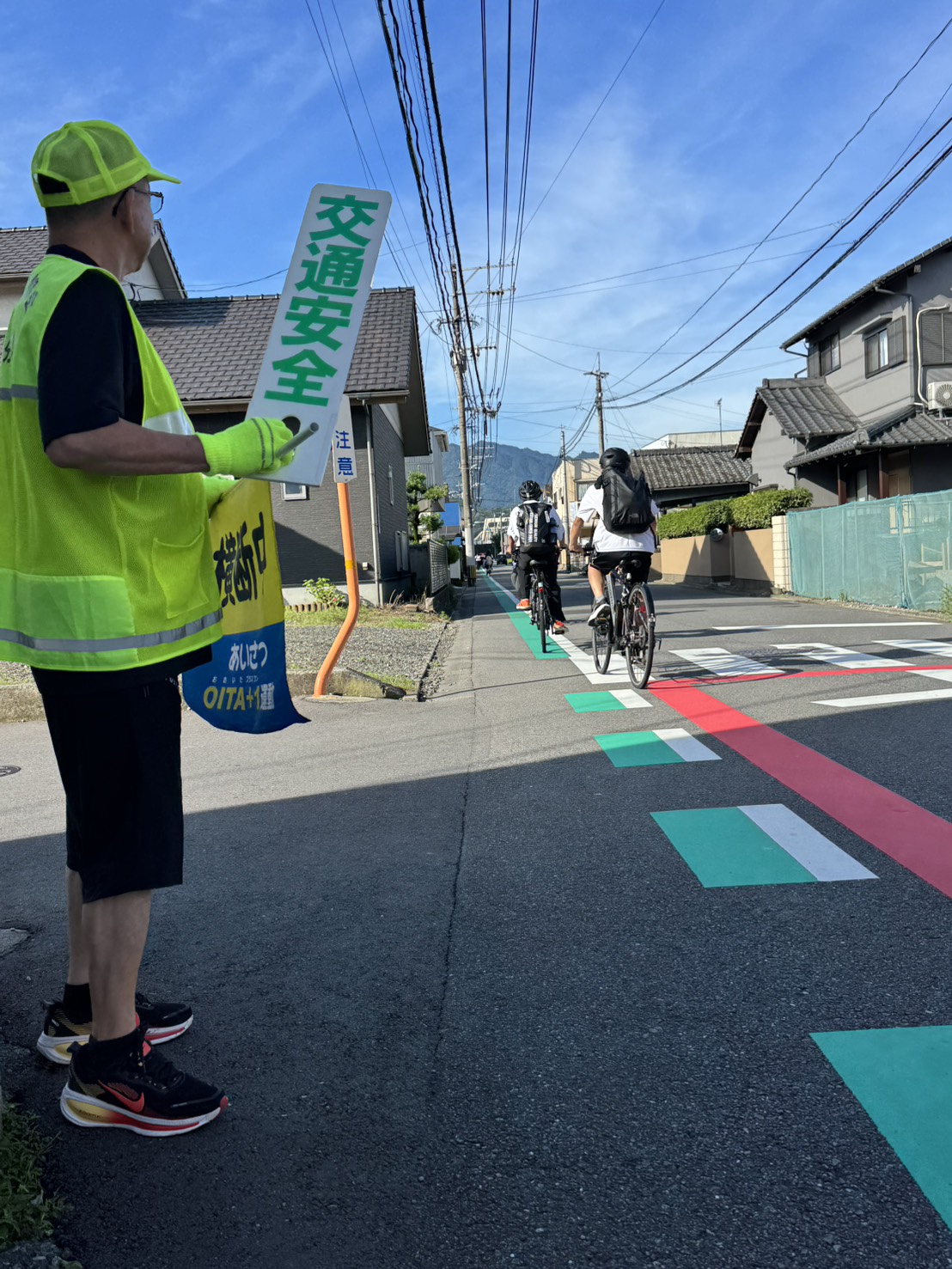 住宅街の道路脇で、黄色い安全ベストと帽子を着用した誘導員が「交通安全」と書かれた標識を持ち、通行する自転車や歩行者を見守っている様子を写した写真。背景には家並みや青空、遠くの山が見え、朝の交通誘導の穏やかな雰囲気が伝わる構図。