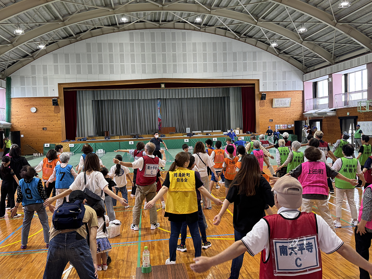 体育館内で、多くの参加者が色分けされたベストを着用し、ステージ前に整列して体を動かしている様子を写した写真。高いアーチ状の天井と木の床の広い空間で、住民の健康増進と交流を目的とした「スマイルボウリング大会」のにぎやかな雰囲気が伝わる構図。