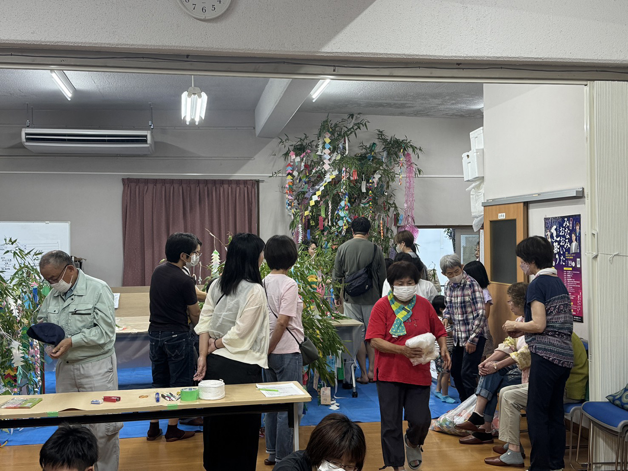 上田町集会所の室内で、笹に色とりどりの短冊や飾りをつけながら作業する住民たちの様子を写した写真。テーブルには紙やテープなどの材料が並び、参加者がマスクを着用して協力しながら七夕飾りを仕上げている。蛍光灯の明るい室内で、地域の恒例行事としての温かい雰囲気が伝わる構図。