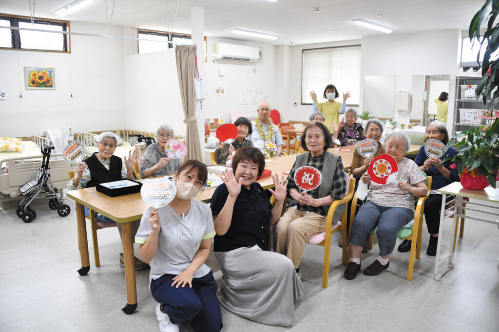 明るい室内で、利用者とスタッフがテーブル周りに集まり、色とりどりの団扇を手に笑顔でポーズを取っている写真。前方にはスタッフがひざ立ちで手を振り、周囲の高齢者も「HAPPY!」などの文字が描かれた団扇を掲げている。背景にはベッドやひまわりの絵があり、デイサービスの和やかな交流風景が伝わる構図。