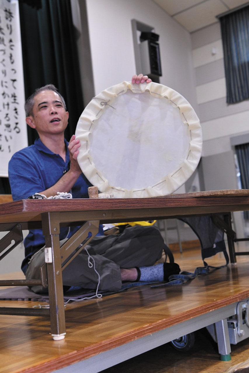 室内のステージ上で、和太鼓の胴を示しながら説明する宮内礼治さんが座っている写真。前方の机には紐や道具が置かれ、背後には縦長の案内幕が立てられている。鹿児島県で和太鼓の製造・販売を手掛ける宮内さんが、南大分公民館の人権講演会で実演を交えながら話している様子が伝わる構図。