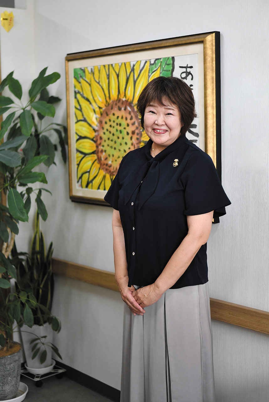 室内の壁に大きなひまわりの絵が飾られ、その前に介護福祉士の福田恵美さんが立っている写真。黒いリボン付きのブラウスと淡いグレーのスカートを着用し、周囲には観葉植物が置かれている。明るい室内で、デイサービスわくわくおひさまランド・さんさんおひさまランドの穏やかな雰囲気が伝わる構図。