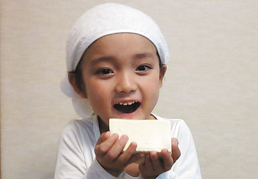 豆腐を持って笑顔の子どもの写真。頭に白い布をかぶり髪をまとめ白いシャツの清潔感のある服装。