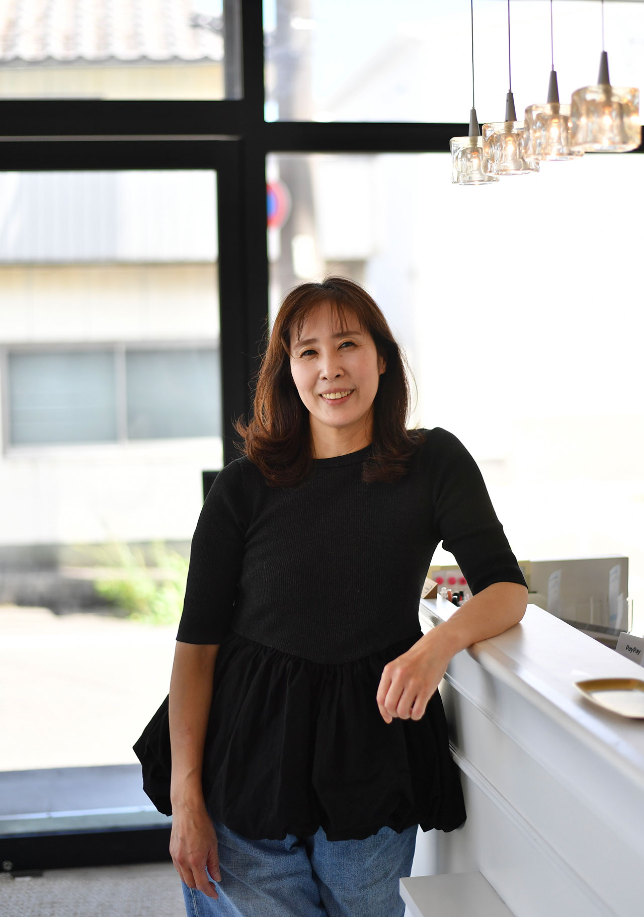 白いカウンターの前に立つ美容師・牧野朋美さんの写真。黒いトップスとジーンズを着用し、背後には大きな窓から自然光が差し込む明るい室内が広がっている。天井にはガラスのペンダントライトが並び、落ち着いた雰囲気の美容室空間が伝わる構図。